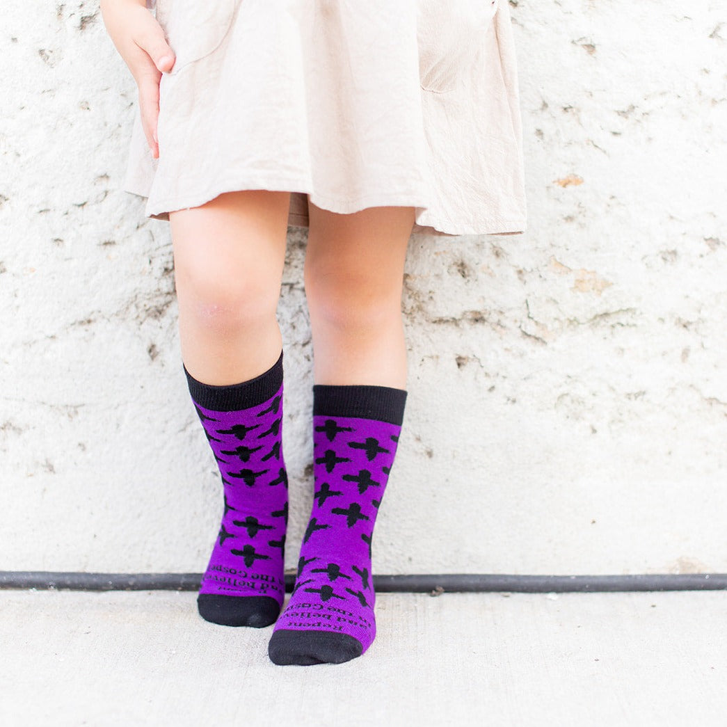 girl standing against a wall wearing Purple socks with black ash crosses that say "repent and believe in the gospel' across the toe